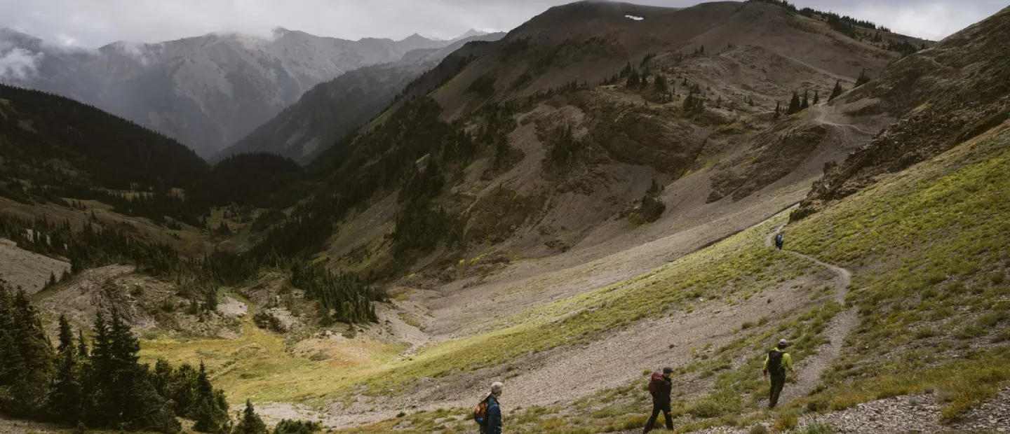 Three hikers walk along a winding trail through a mountainous landscape under a cloudy sky.