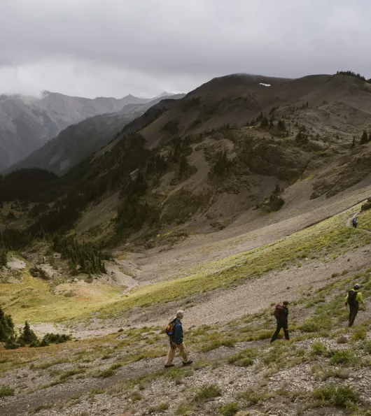 Three hikers walk along a winding trail through a mountainous landscape under a cloudy sky.